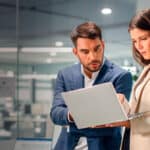Homem e mulher em um ambiente corporativo com um notebook em mãos olhando para a tela