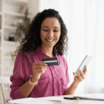 Mulher sentada em uma sala segurando um cartão com uma das mãos e um celular com a outra mão.