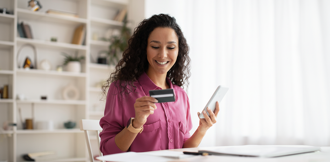 Mulher sentada em uma sala segurando um cartão com uma das mãos e um celular com a outra mão.