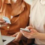 Duas mulheres usando celular e notebook enquanto se atentam à prevenção de fraudes e Machine Learning.
